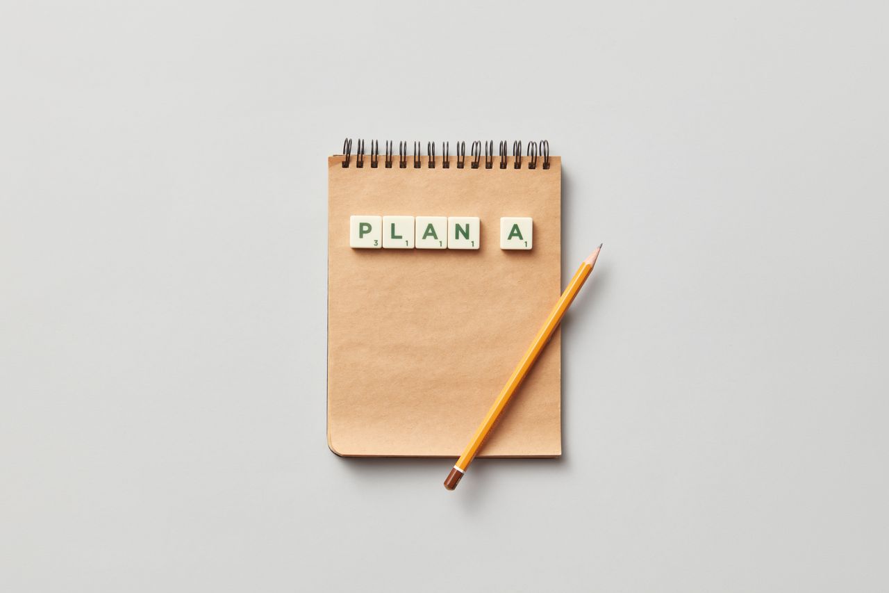 Top view of a notepad with a pencil and scrabble tiles spelling 'PLAN A' on a gray background.